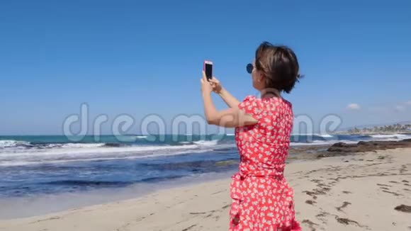 白人妇女拿着相机电话拍着海上风暴站在海滩上的照片她穿着红裙子戴着纱丽视频的预览图