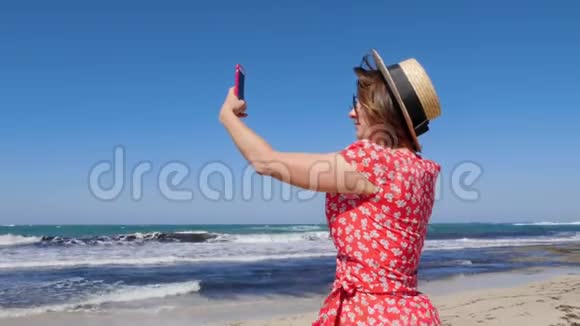 年轻的女人拿着她的手机为狂风暴雨的大海和海浪拍照她戴着帽子穿着带太阳镜的红色连衣裙视频的预览图