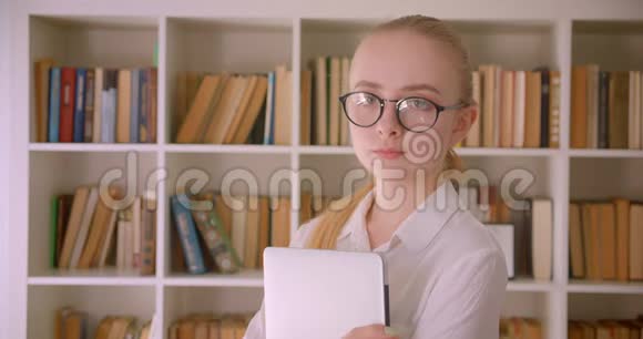 年轻漂亮的白种人金发女学生戴着眼镜拿着笔记本电脑看着镜头微笑的特写照片视频的预览图