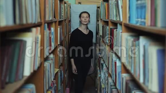 美丽的年轻女孩留着长长的黑发走到图书馆的垂直书架之间微笑着视频的预览图