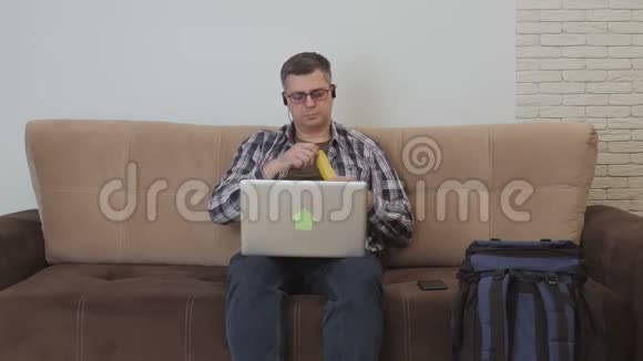 一个中年男人坐在沙发上耳朵里戴着耳机他喜欢笔记本电脑吃香蕉他旁边是一个视频的预览图