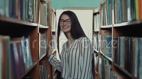 美丽的年轻女孩留着长长的黑发走到图书馆的垂直书架之间微笑着视频的预览图