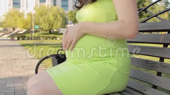 一位孕妇坐在公园的长凳上从下到上抚摸她的腹部怀孕期间在大自然中行走等待视频的预览图