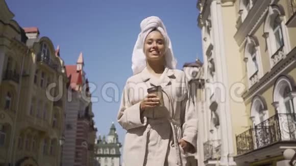 美丽的微笑年轻女子穿着浴袍头上戴着毛巾走在街上喝着咖啡自信的女孩视频的预览图