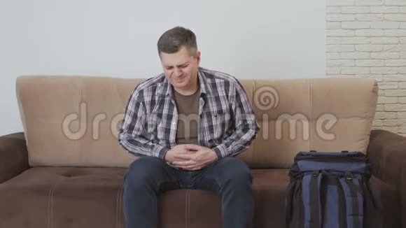 一个中年男人坐在沙发上抱着他的胃痛苦地扭动着沙发旁边是一个大型旅游背包视频的预览图