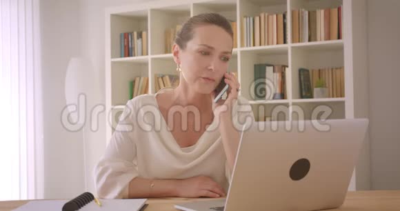 用笔记本电脑和电话交谈的老白种人黑发女商人的特写照片视频的预览图