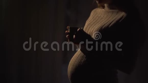 微笑着年轻的孕妇站在温暖的毛衣在黑暗的房间里喝热茶女士在等视频的预览图