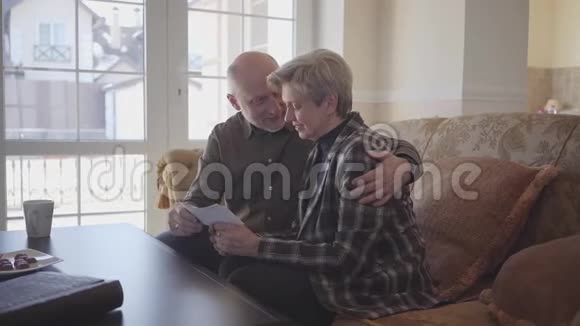 老两口坐在沙发上看着老照片男人拥抱女人用爱和激情看着她令人惊叹视频的预览图