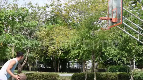 亚洲男人手里拿着篮球跳着跳着扔篮圈公园里的背景树视频的预览图