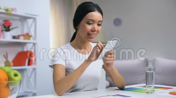 迷人的微笑女性通过智能手机应用程序聊天坐在家里的桌子上视频的预览图