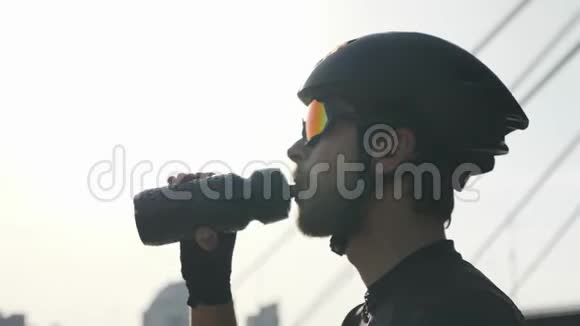 自信的骑自行车的人带着红胡子从瓶子里喝水戴着头盔和太阳镜桥和河的背景视频的预览图