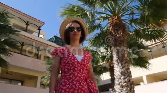 年轻迷人的女人享受阳光戴着帽子和太阳镜背景上有棕榈树花园酒店视频的预览图