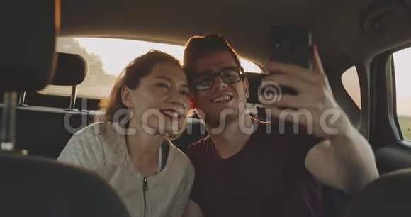 穿着毛衣和帽子的漂亮夫妇拍自拍照片坐在森林路边的汽车后备箱里年轻家庭视频的预览图
