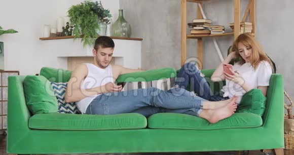 男人和女人坐在家里的绿色沙发上浏览手机视频的预览图