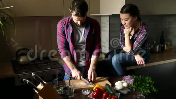 漂亮的年轻夫妇在家厨房做饭时吃健康的食物男人是切沙拉女人坐在桌子上看着场景f视频的预览图
