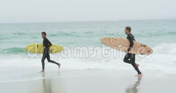 两名男性冲浪者在海滩上用冲浪板跑步的侧视4k视频的预览图
