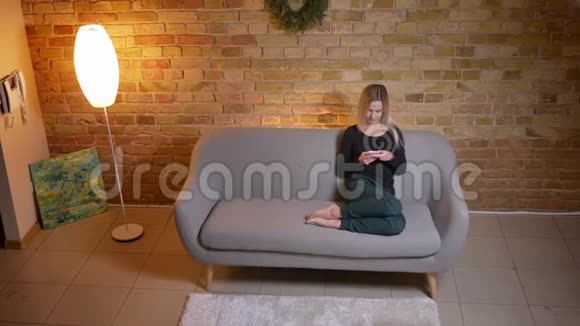 在舒适的室内沙发上一位年轻漂亮的金发女性使用手机坐在沙发上的特写镜头视频的预览图