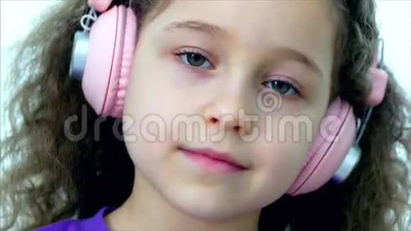 一个卷发可爱的孩子的肖像一个穿着紫色t恤戴着粉色耳机听着的白种人小女孩视频的预览图