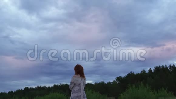 年轻的女孩子穿着白色的裙子站在前景享受着罕见的发光的自然天空喜怒无常的寒冷日落视频的预览图
