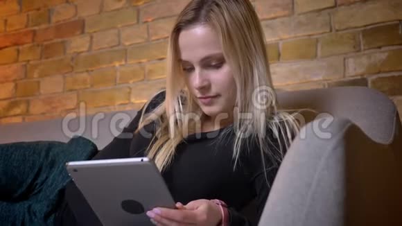 年轻漂亮的金发女性使用平板电脑躺在沙发上悠闲地躺在舒适的室内视频的预览图