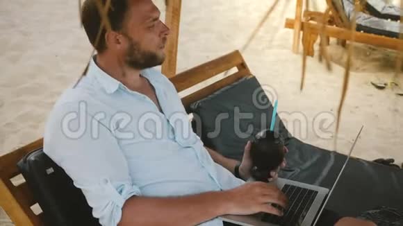 成功的严肃中年商人坐在海滩上的躺椅上带着水果饮料和笔记本电脑思考视频的预览图