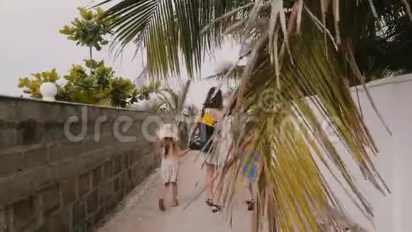 快乐的年轻女子和小男孩一起走着小女孩背着袋子沿着小棕榈树的小巷走向海边视频的预览图