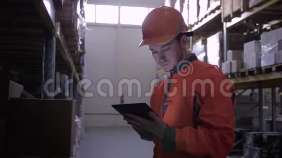 年轻的仓库工人戴着橙色安全帽靠近大架带着货物使用平板电脑视频的预览图
