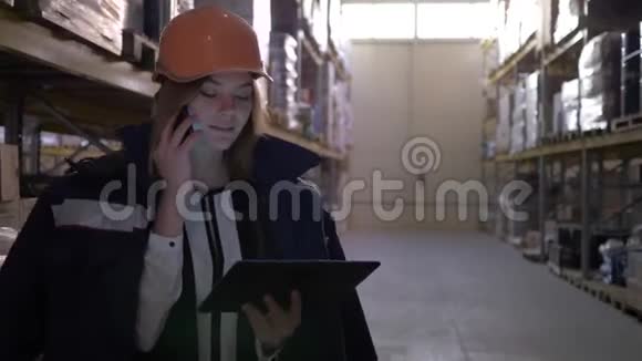 仓库里戴着橙色头盔的年轻女子在用手机和平板电脑说话视频的预览图