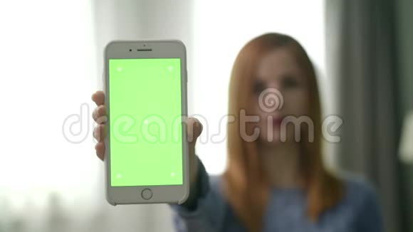 房间里漂亮的年轻女人用绿色屏幕展示智能手机视频的预览图