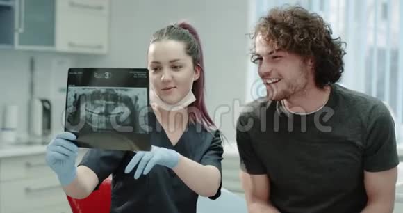 在一家牙科诊所戴着蓝色手套的年轻牙医向一位年轻的病人解释了牙齿X光的结果他们有一个视频的预览图