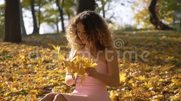 中弹微笑的卷曲女孩坐在公园的草地上编织着花环温暖的秋日柔和的灯光4K视频的预览图