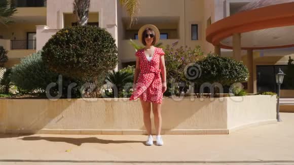 年轻快乐的白种人女人走在酒店花园的桥上走向镜头穿着红色的连衣裙戴着帽子和眼镜慢慢视频的预览图