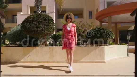 年轻快乐的白种人女人走在酒店花园的桥上对着镜头穿着红色的连衣裙帽子和眼镜视频的预览图