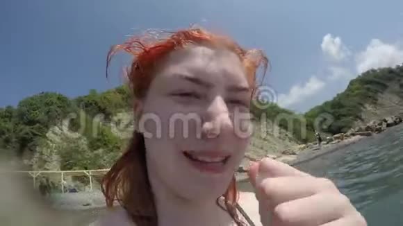 白种人快乐有趣的红发女孩看着镜头背景的海洋和沙滩陡峭的绿色海滩视频的预览图