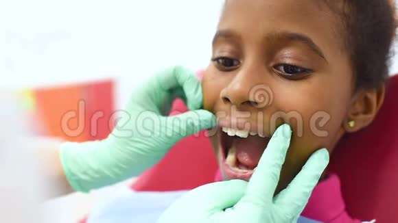 可爱的深色皮肤的非裔美国女孩坐在一个红色的黄色儿童牙科椅上看牙医视频的预览图