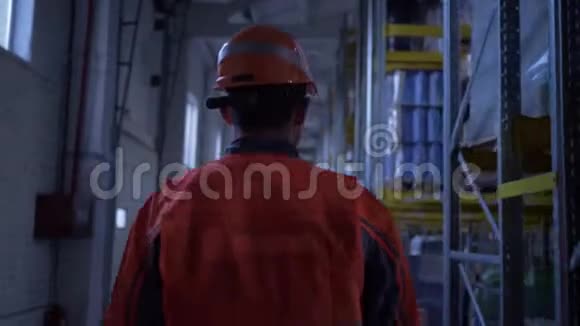 身穿橙色头盔和制服的年轻人穿过大仓库用箱子检查货架视频的预览图