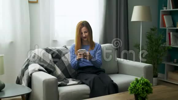 年轻开朗的女孩坐在沙发上舒适的客厅里有智能手机视频的预览图