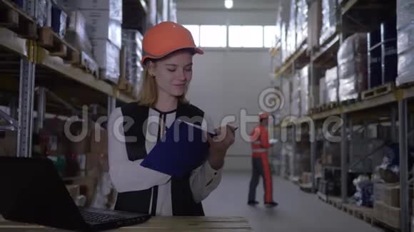 女员工戴安全帽的肖像与笔记本电脑一起工作并在仓库工人的背景下在记事本上做笔记视频的预览图