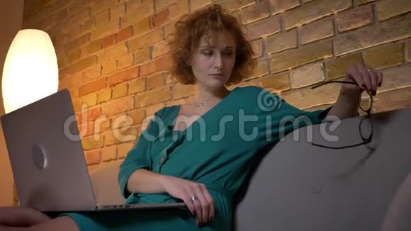 姜黄色卷发的白种人女孩坐在笔记本电脑前在舒适的家里戴眼镜的肖像视频的预览图