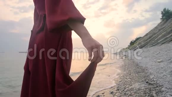 一个年轻女孩的手特写她穿着一件红色的裙子从身后走在日落时海边的岩石海滩上A视频的预览图