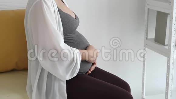 孕妇在家卧室靠近窗户的沙发上用手轻轻地摸着肚子人民的生活方式和视频的预览图