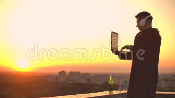 日落时分一个戴着耳机的自由职业者站在屋顶上写着键盘代码页小生意听着听着视频的预览图
