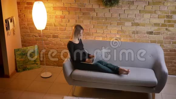 在笔记本电脑上在线学习的年轻漂亮女学生坐在室内沙发上的侧视图视频的预览图