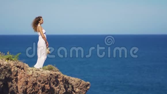 穿着白色裙子的女孩站在岩石悬崖上一个女人望着远处一望无际的海面视频的预览图