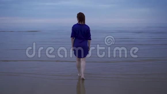 年轻的红发女子穿着蓝色连衣裙湿发在水面上行走溅起水花忧郁而寒冷的日落视频的预览图