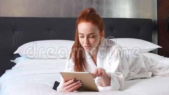年轻的红头发妇女使用平板电脑躺在酒店房间的床上视频的预览图