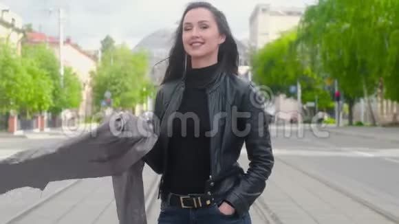 穿着黑色皮夹克和灰色围巾的年轻女孩站在一条城市街道的背景上她笑了视频的预览图