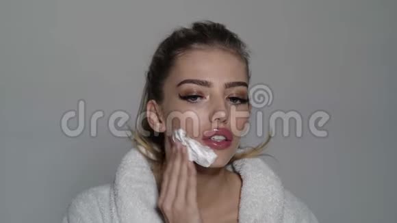 女孩在忙碌的脸上穿着浴袍用泡沫覆盖脸用于剃须灰色背景一个满脸泡沫的女人视频的预览图