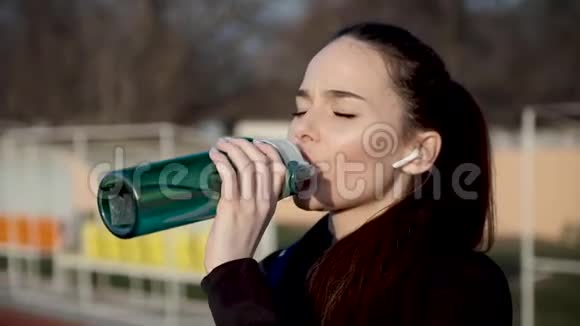 在体育场戴无线耳机的女孩从一个特殊的运动瓶里喝水视频的预览图