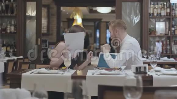 肖像可爱的年轻女子坐在餐厅的桌子上有一个自信的留着胡子的男人给她看视频的预览图